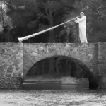 Solénoïde - Mission 225 : photo de Stephan Micus sur un pont avec son instrument, une longue trompette tibétaine appelée dung chen.