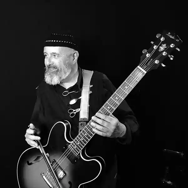 Photo de l'artiste Serge Pesce avec sa guitare accommodée. L'artiste présente et associe différents accessoires à sa guitare : archet, ciseaux, cuillère.